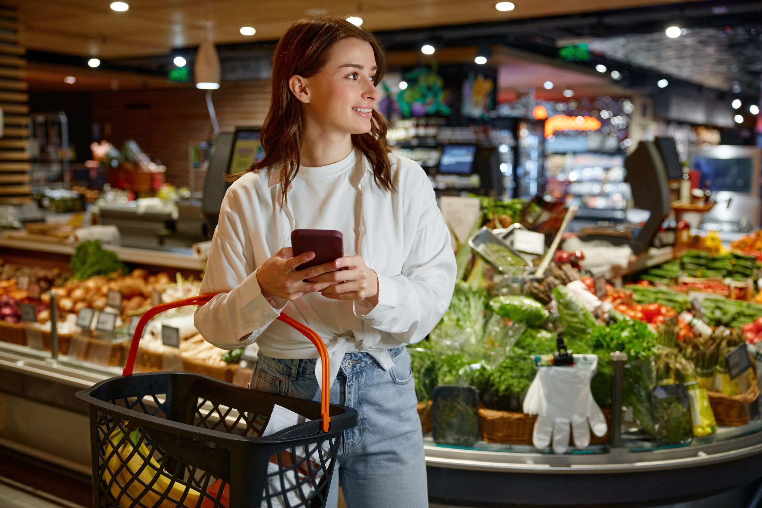 Woman in a white shirt and jeans smiles while holding a smartphone in a supermarket produce section, with a large shopping basket in front of her.